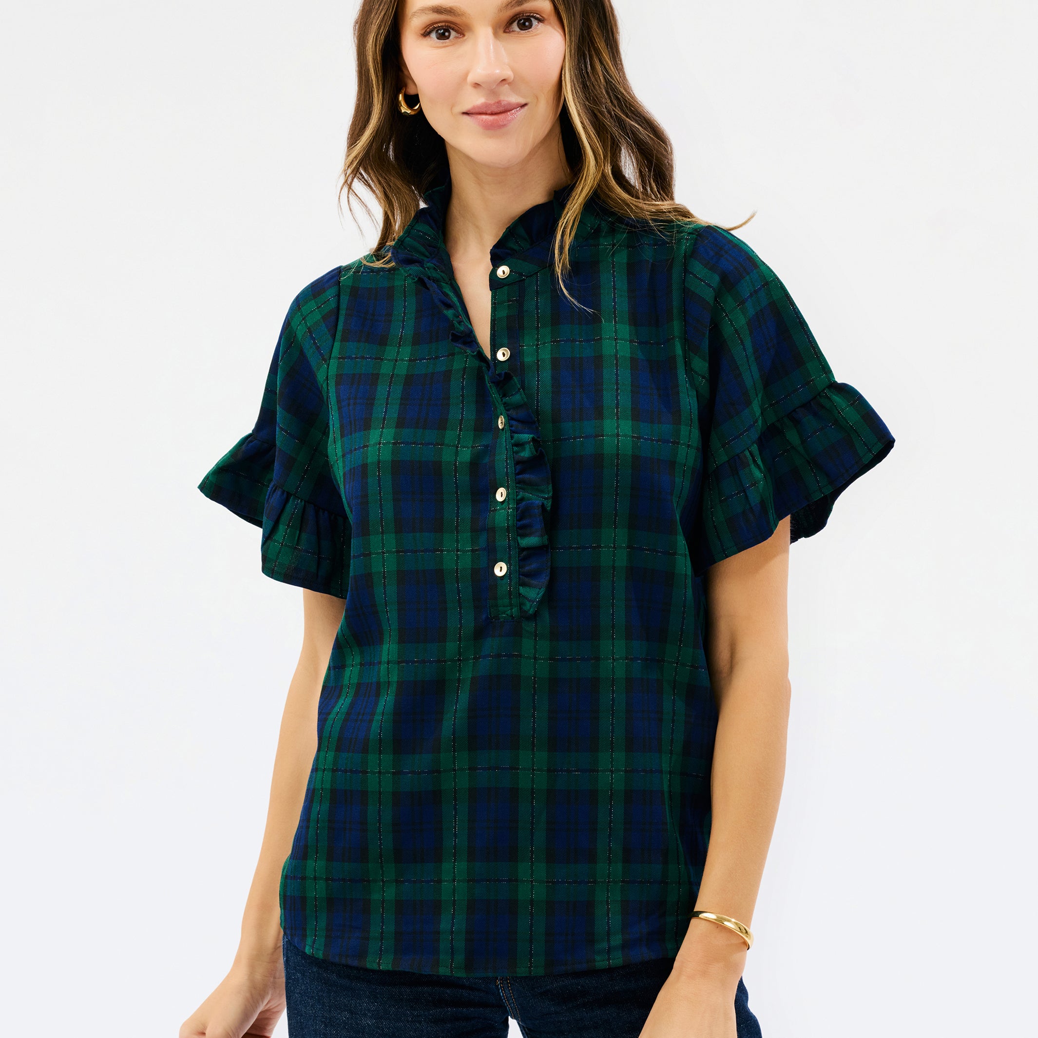 A woman modeling a short-sleeve button-front plaid blouse in dark green and navy with ruffled trim at the collar and sleeve cuffs, paired with dark jeans against a light gray studio background.