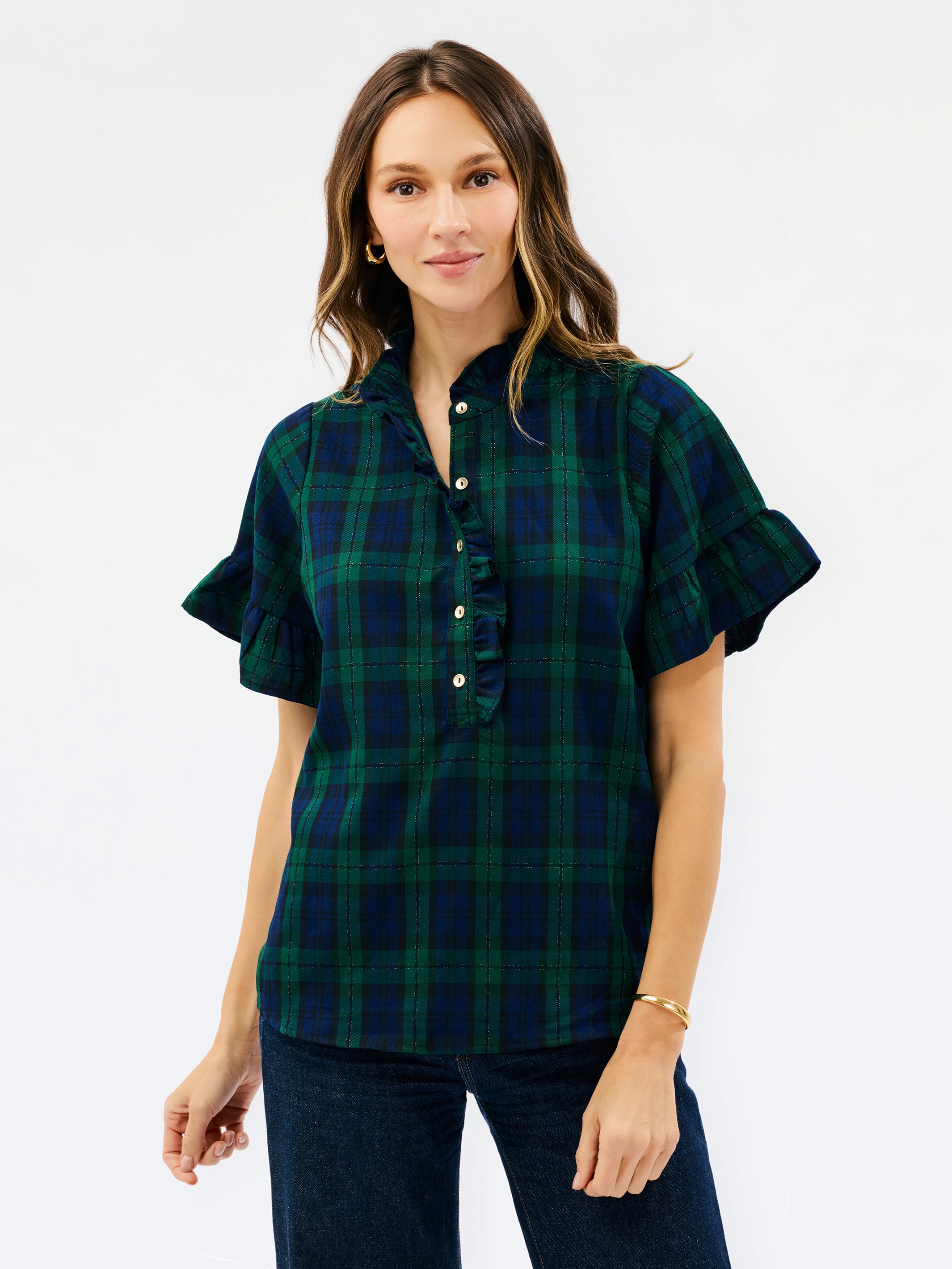 A woman modeling a short-sleeve button-front plaid blouse in dark green and navy with ruffled trim at the collar and sleeve cuffs, paired with dark jeans against a light gray studio background.