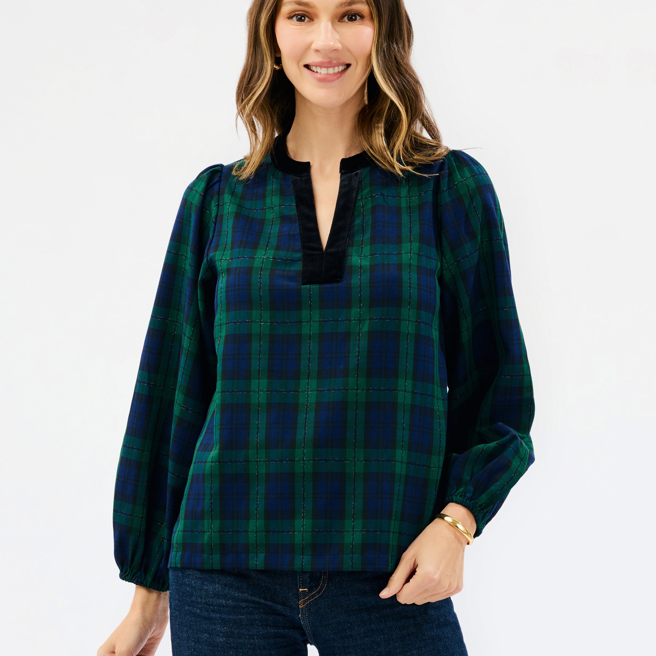 A woman wearing a long-sleeve plaid blouse in dark green and navy with a black split neckline, paired with dark blue jeans, against a plain white studio background.