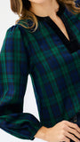 A close-up of a woman wearing a long-sleeve plaid blouse in dark green and navy blue with a black velvet V-neckline trim, paired with dark jeans.