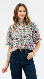 A woman modeling a short-sleeve button-up blouse with a dense floral and botanical print in green, burgundy, and cream, paired with dark blue jeans against a plain white studio background.
