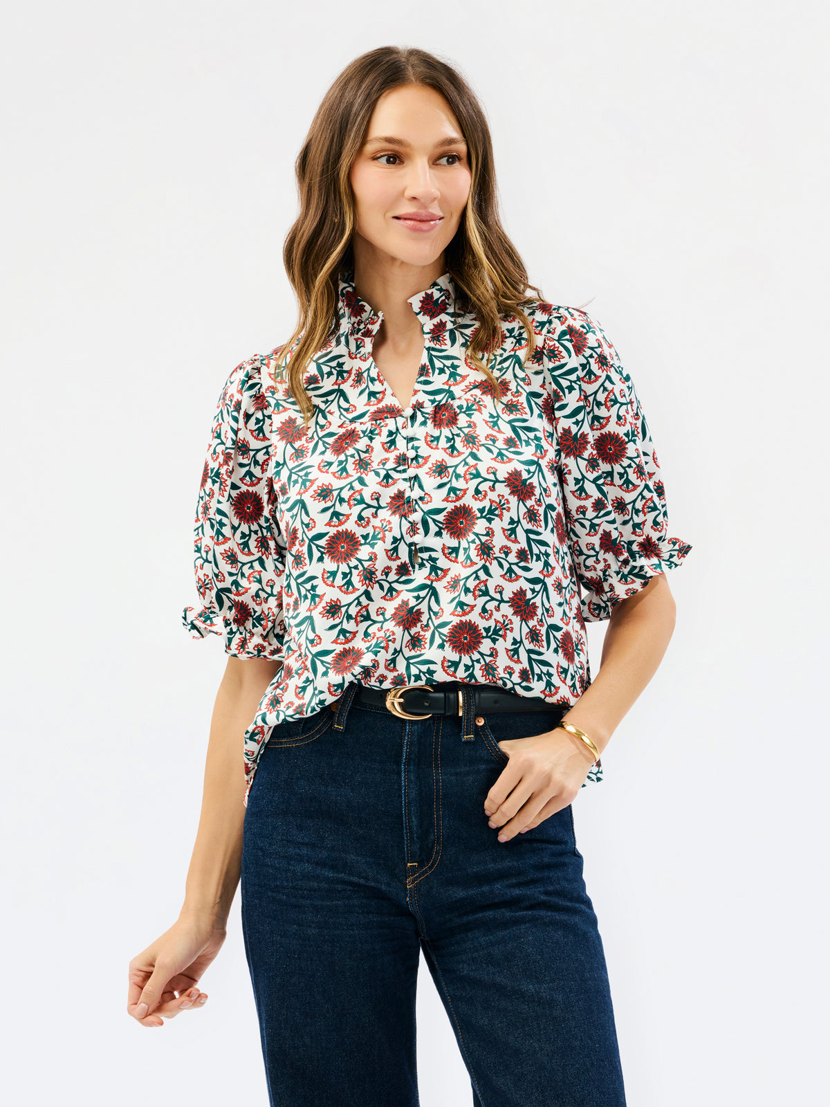 A woman modeling a short-sleeve button-up blouse with a dense floral and botanical print in green, burgundy, and cream, paired with dark blue jeans against a plain white studio background.