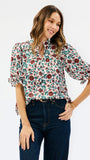 A woman modeling a short-sleeve button-up blouse with a dense floral print in green, burgundy, and cream, paired with dark blue jeans against a plain white studio background.