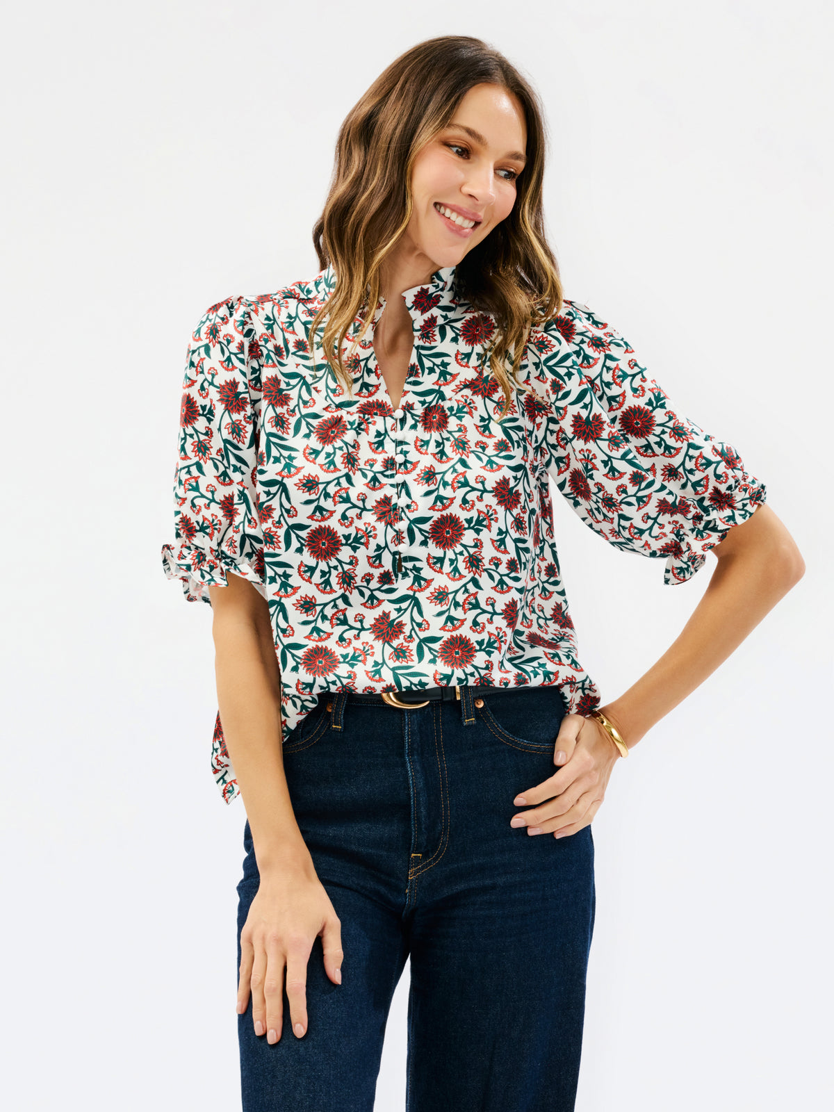 A woman modeling a short-sleeve button-up blouse with a dense floral print in green, burgundy, and cream, paired with dark blue jeans against a plain white studio background.