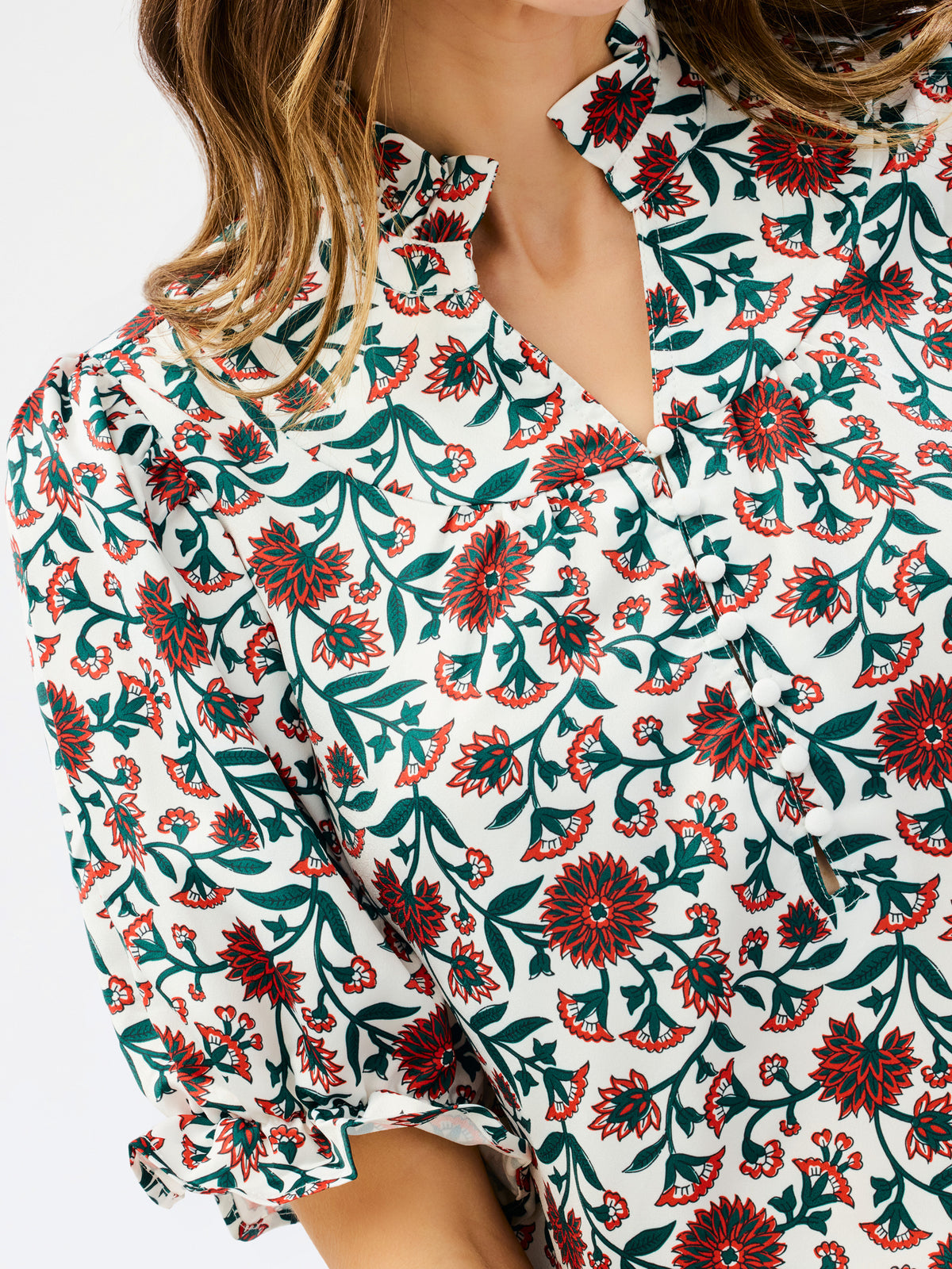 A close-up of a woman wearing a short-sleeve blouse with a white base and an allover floral print in dark green and red-orange.