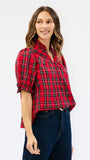 A woman modeling a red and black plaid short-sleeve button-up shirt with dark blue jeans, photographed against a plain white studio background.
