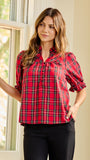 A woman wearing a red and black plaid button-up blouse with puff sleeves, standing indoors near a window.