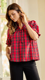 A woman wearing a red and black plaid short-sleeve blouse with puff sleeves, paired with black pants, standing in a brightly lit room with white columns.