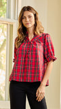 A woman wearing a red and black plaid short-sleeve blouse with puff sleeves and a tie-neck detail, paired with black pants, standing in a brightly lit room with a window.