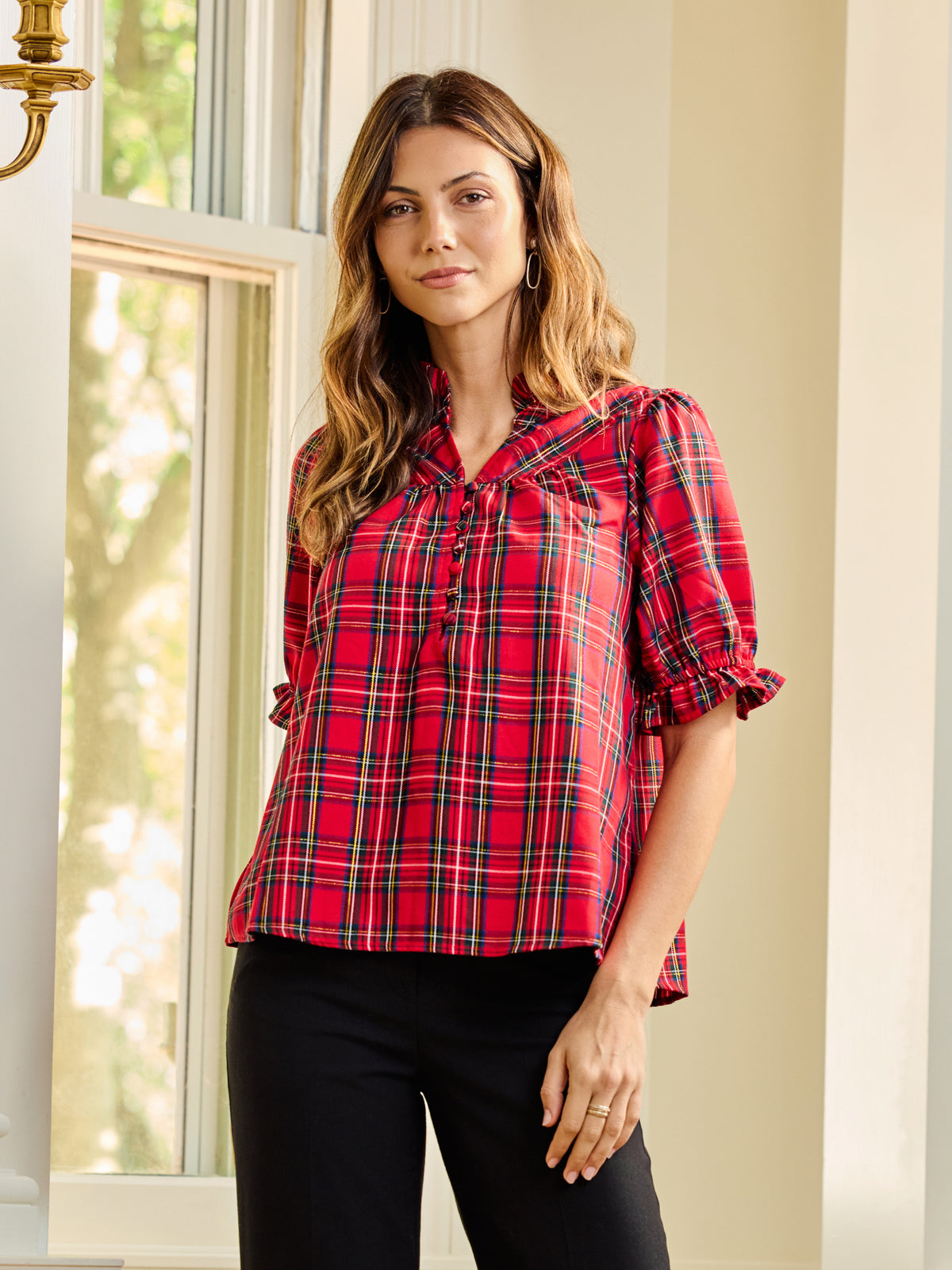 A woman wearing a red and black plaid short-sleeve blouse with puff sleeves and a tie-neck detail, paired with black pants, standing in a brightly lit room with a window.