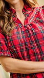 A close-up of a woman wearing a red plaid blouse with puff sleeves and a ruffled collar. The blouse features a button placket and a tartan pattern with red, green, and yellow lines.