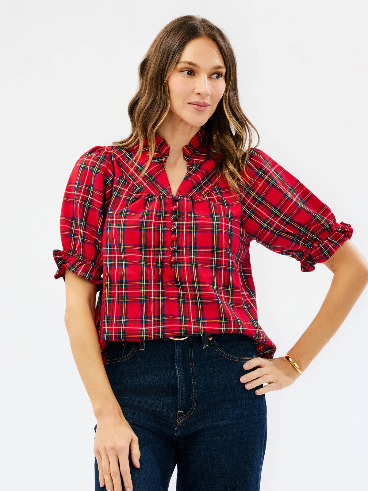 A woman wearing a red and black plaid short-sleeve blouse with puff sleeves and dark blue jeans, standing against a plain white studio background.