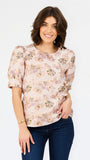 A woman modeling a light pink short-sleeve blouse with a multicolor floral pattern, paired with dark blue jeans, against a plain white background.