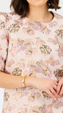 A close-up of a woman wearing a short-sleeve blouse with a soft blush pink base and an allover floral print in lavender, beige, and muted green. She is wearing a gold bracelet against a clean white background.