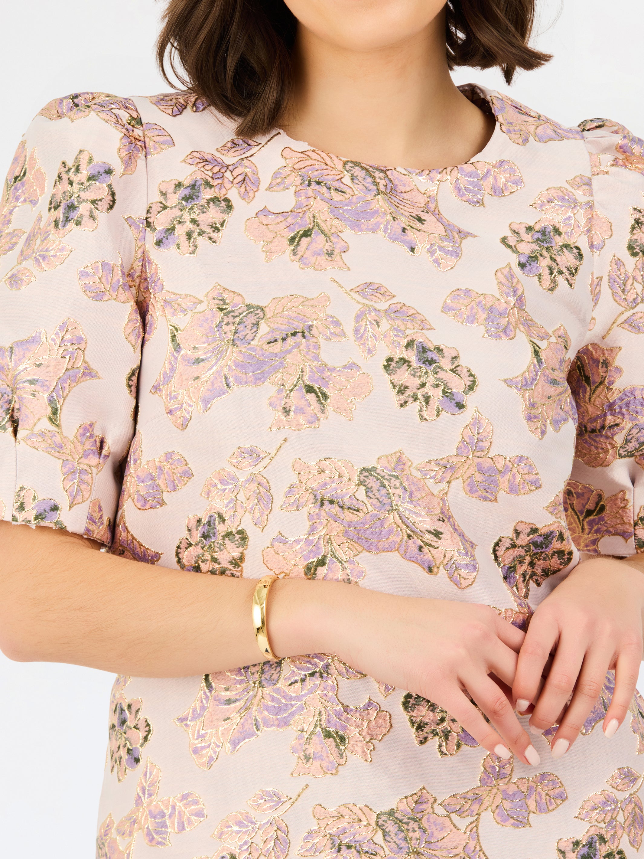 A close-up of a woman wearing a short-sleeve blouse with a soft blush pink base and an allover floral print in lavender, beige, and muted green. She is wearing a gold bracelet against a clean white background.