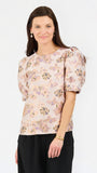 A woman modeling a short-sleeve blouse with puff sleeves and an all-over floral pattern in soft pink, beige, and muted green tones, paired with black trousers, against a plain white studio background.