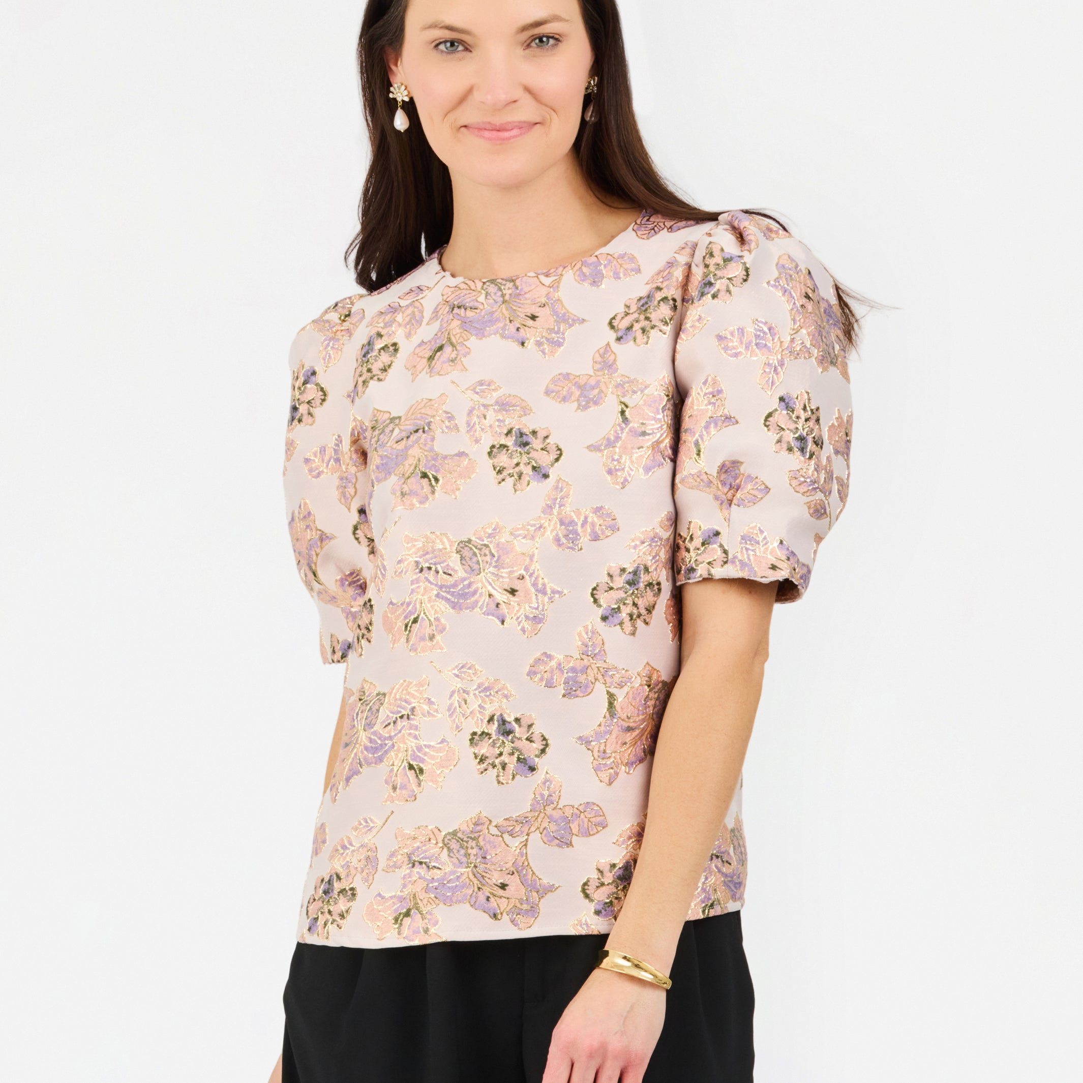 A woman modeling a short-sleeve blouse with puff sleeves and an all-over floral pattern in soft pink, beige, and muted green tones, paired with black trousers, against a plain white studio background.