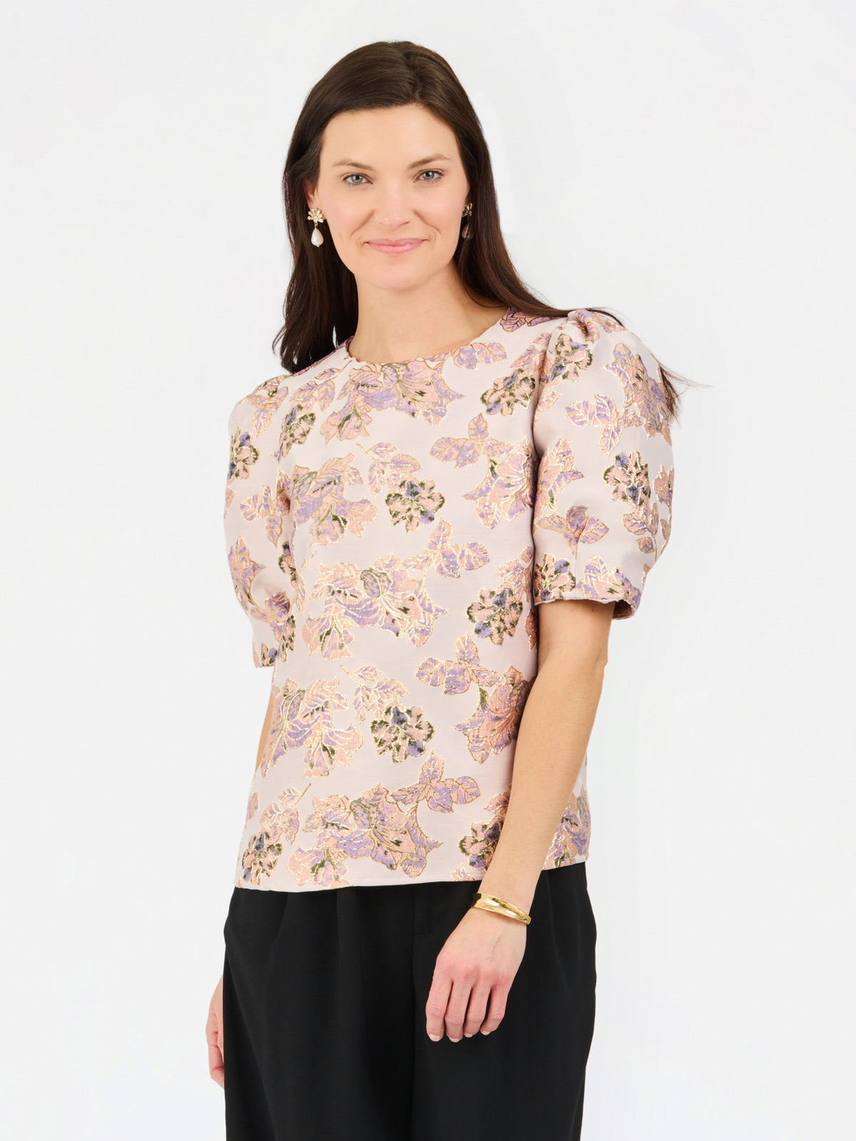A woman modeling a short-sleeve blouse with puff sleeves and an all-over floral pattern in soft pink, beige, and muted green tones, paired with black trousers, against a plain white studio background.