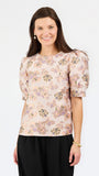 A woman modeling a short-sleeve blouse with puff sleeves and an all-over floral print in soft pink, beige, and purple tones, paired with black trousers, against a plain white studio background.