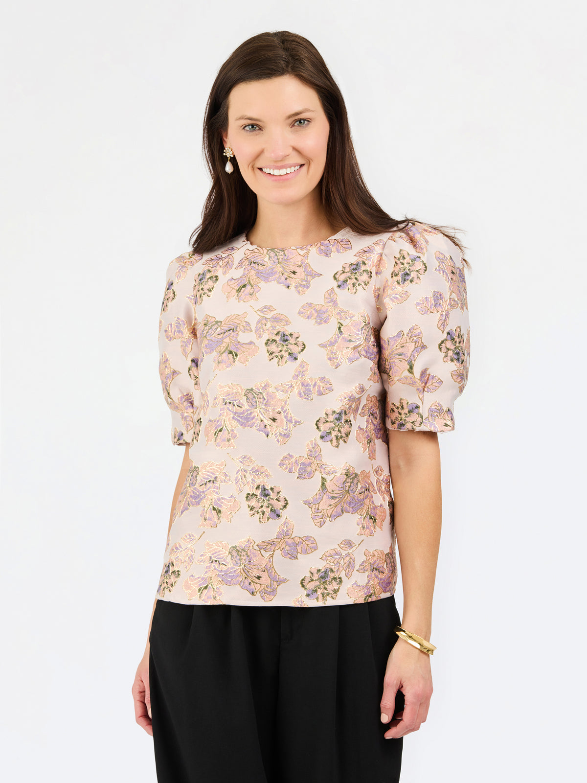 A woman modeling a short-sleeve blouse with puff sleeves and an all-over floral print in soft pink, beige, and purple tones, paired with black trousers, against a plain white studio background.