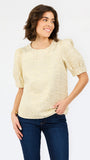 A woman wearing a pale yellow textured short-sleeve blouse with puff sleeves and dark blue jeans, standing against a plain white studio background.