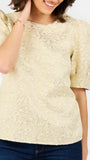 A woman wearing a cream-colored textured short-sleeve top with puff sleeves and dark blue jeans, shown from the waist up against a plain white background.