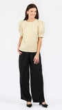 A woman modeling a short-sleeve textured top with puffed shoulders in a light beige color, paired with wide-leg black trousers and black pointed flats, against a plain white studio background.