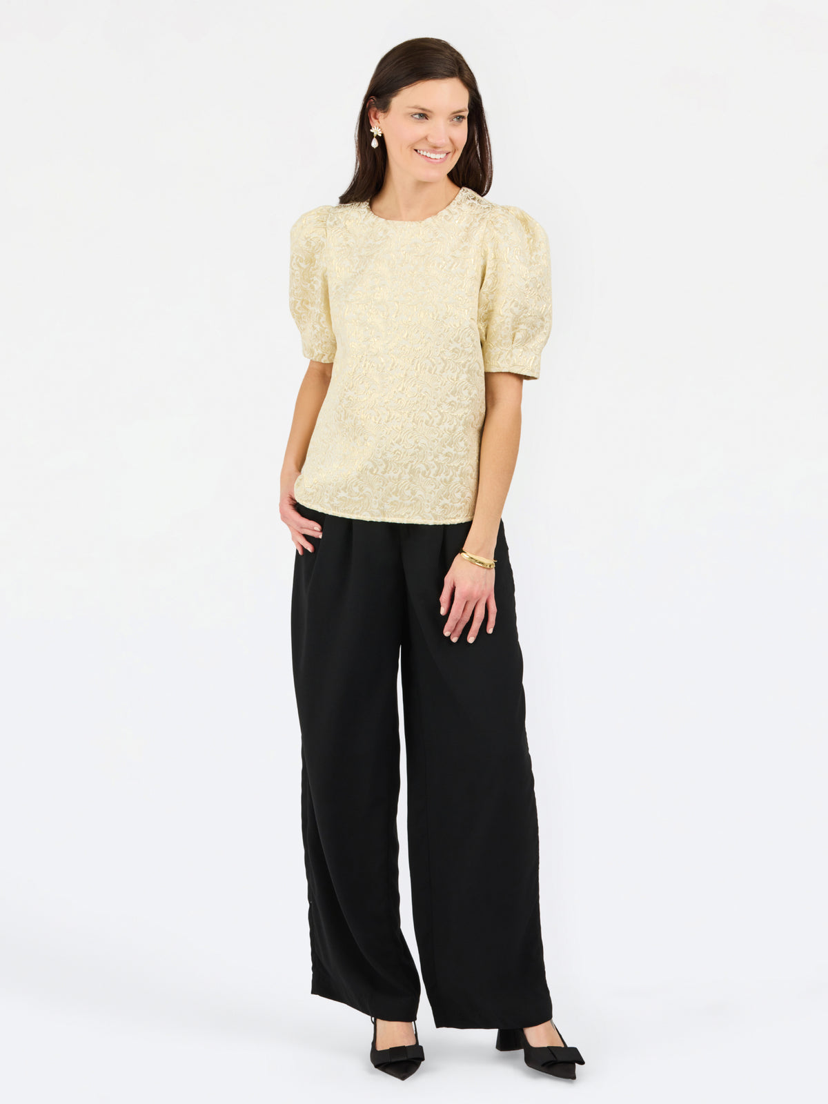 A woman modeling a short-sleeve textured top with puffed shoulders in a light beige color, paired with wide-leg black trousers and black pointed flats, against a plain white studio background.