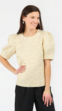 A woman modeling a short-sleeve textured top with puffed sleeves in a light champagne-gold color, paired with black pants, against a plain white studio background.