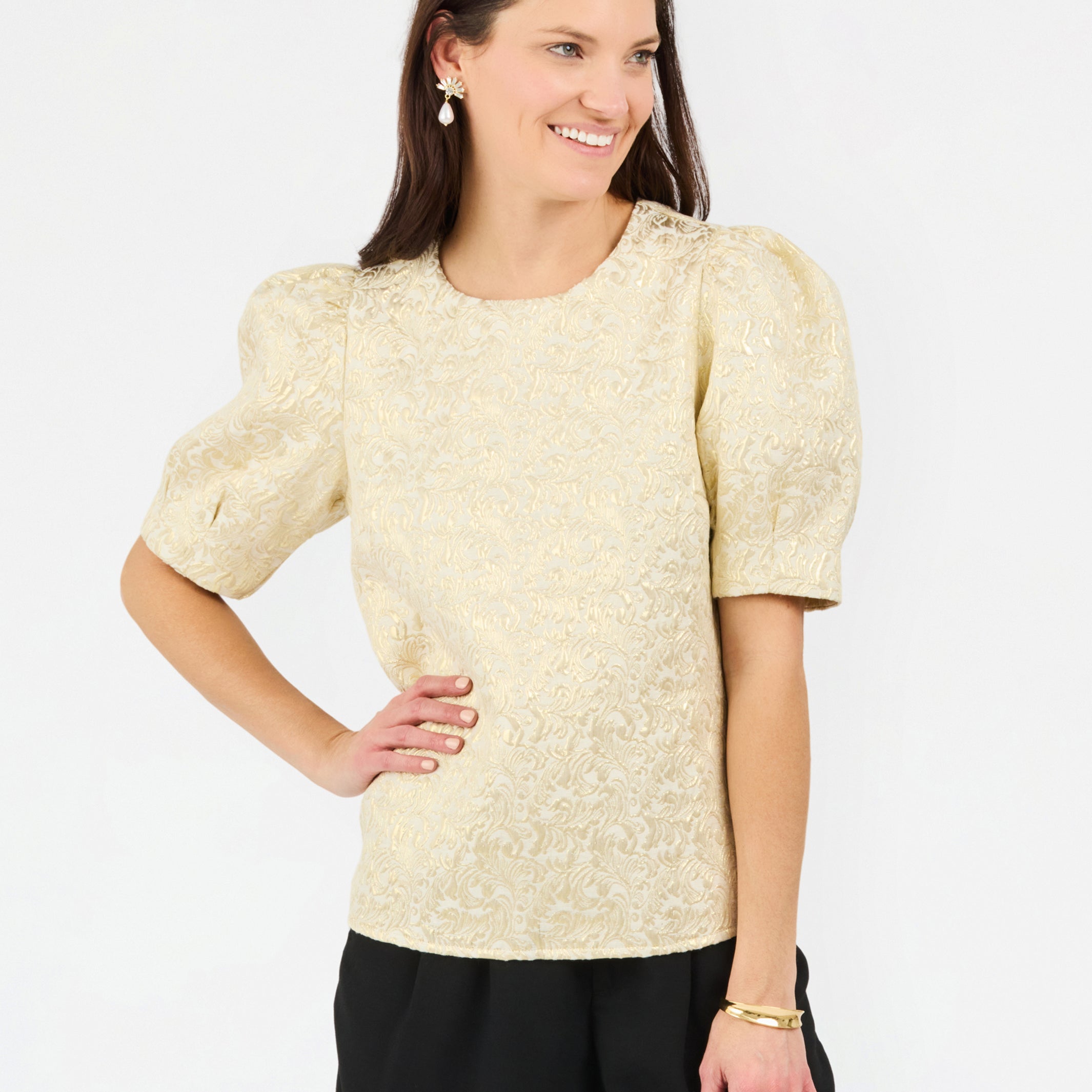 A woman modeling a short-sleeve textured top with puffed sleeves in a light champagne-gold color, paired with black pants, against a plain white studio background.