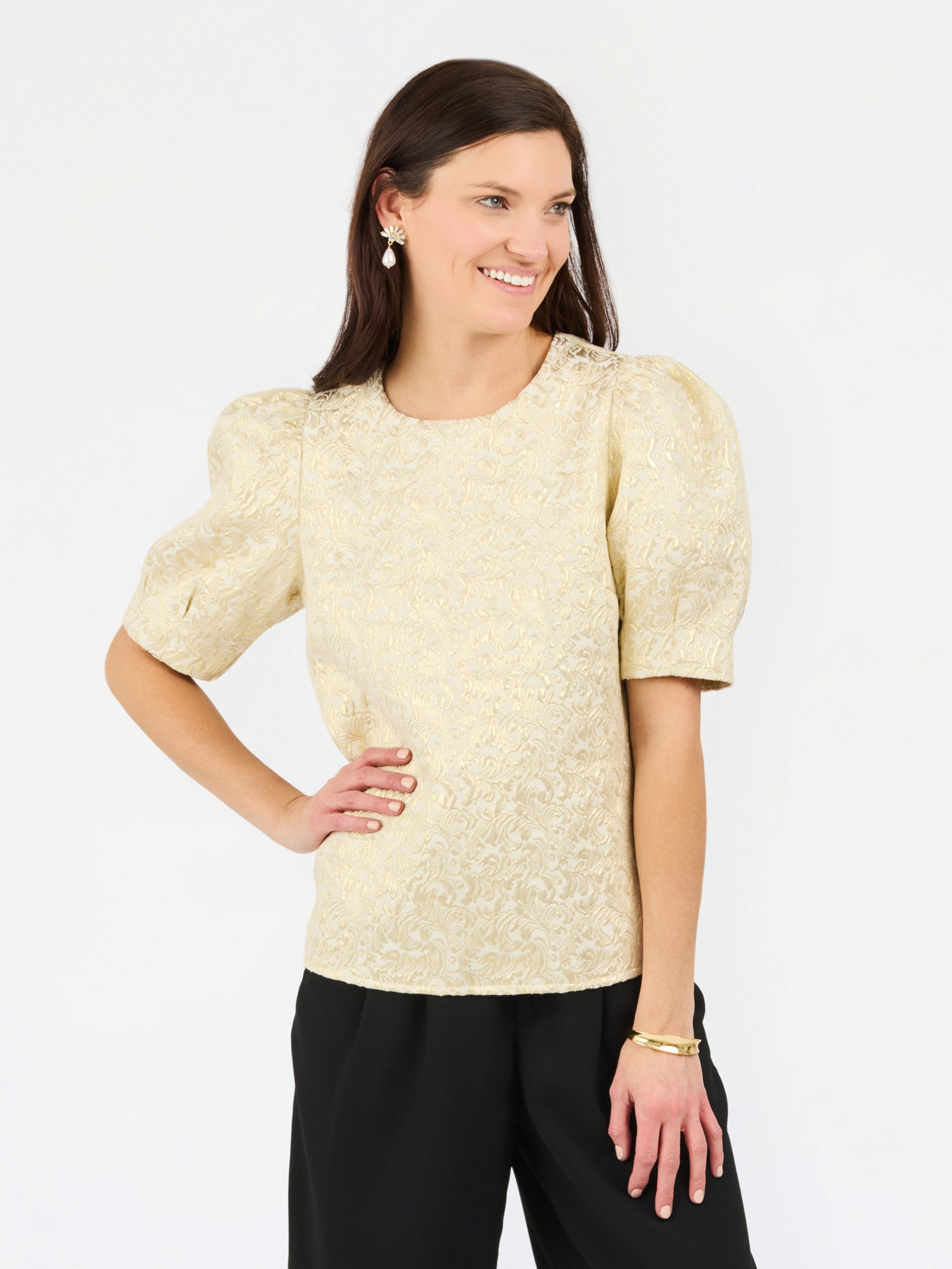 A woman modeling a short-sleeve textured top with puffed sleeves in a light champagne-gold color, paired with black pants, against a plain white studio background.