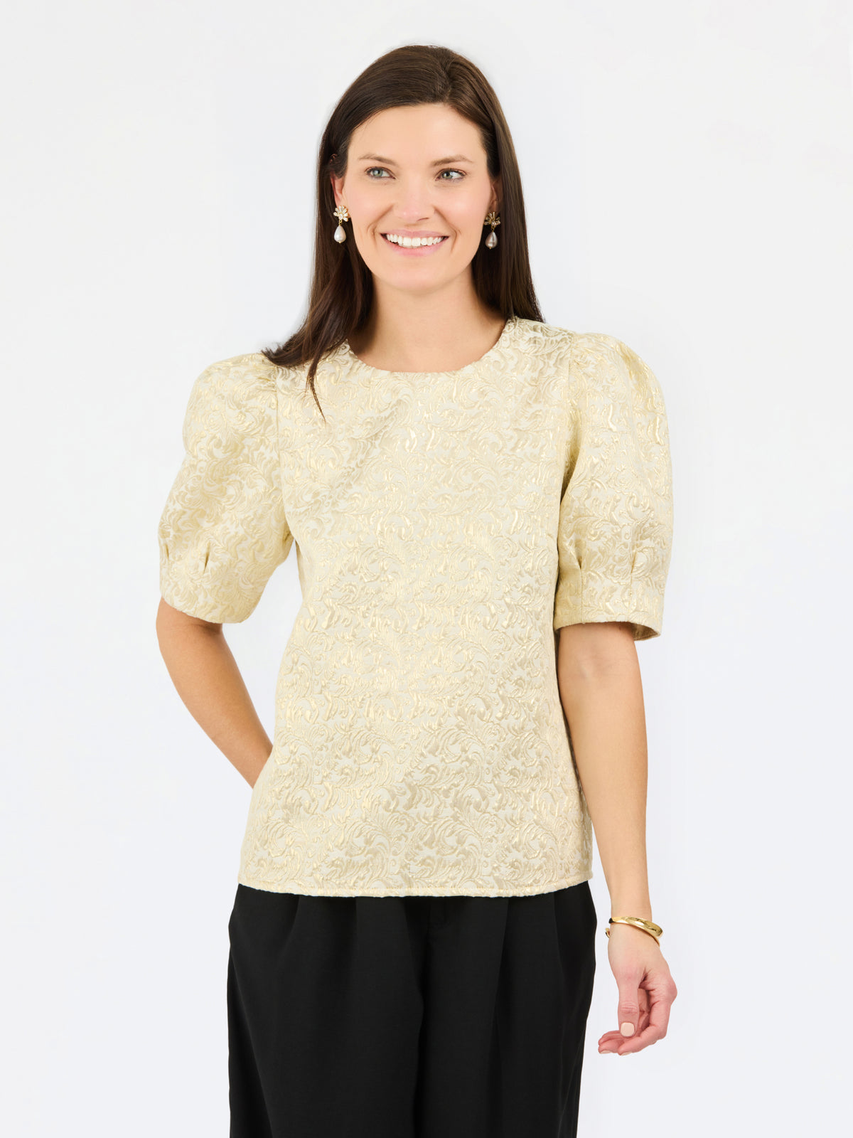 A woman modeling a short-sleeve textured top in a light champagne-gold color, paired with black pants, against a plain white studio background.