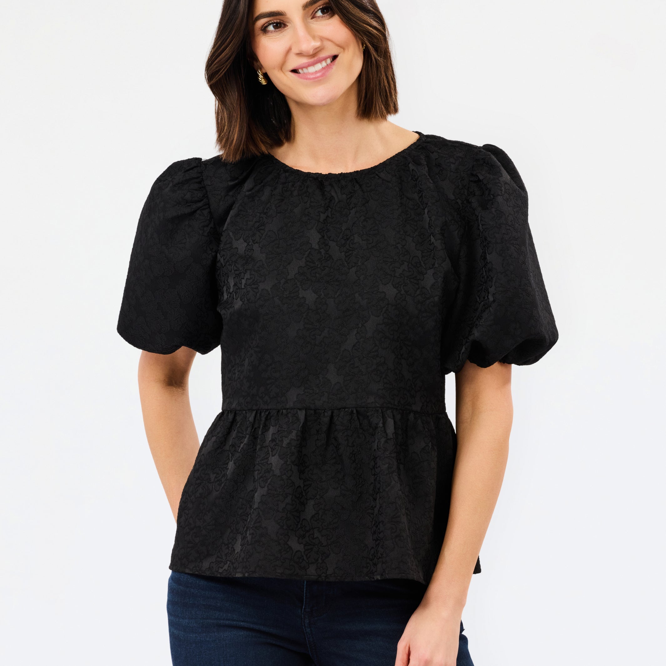 A woman modeling a black short-sleeve blouse with textured puff sleeves and a peplum waist, paired with dark blue jeans, against a plain white studio background.