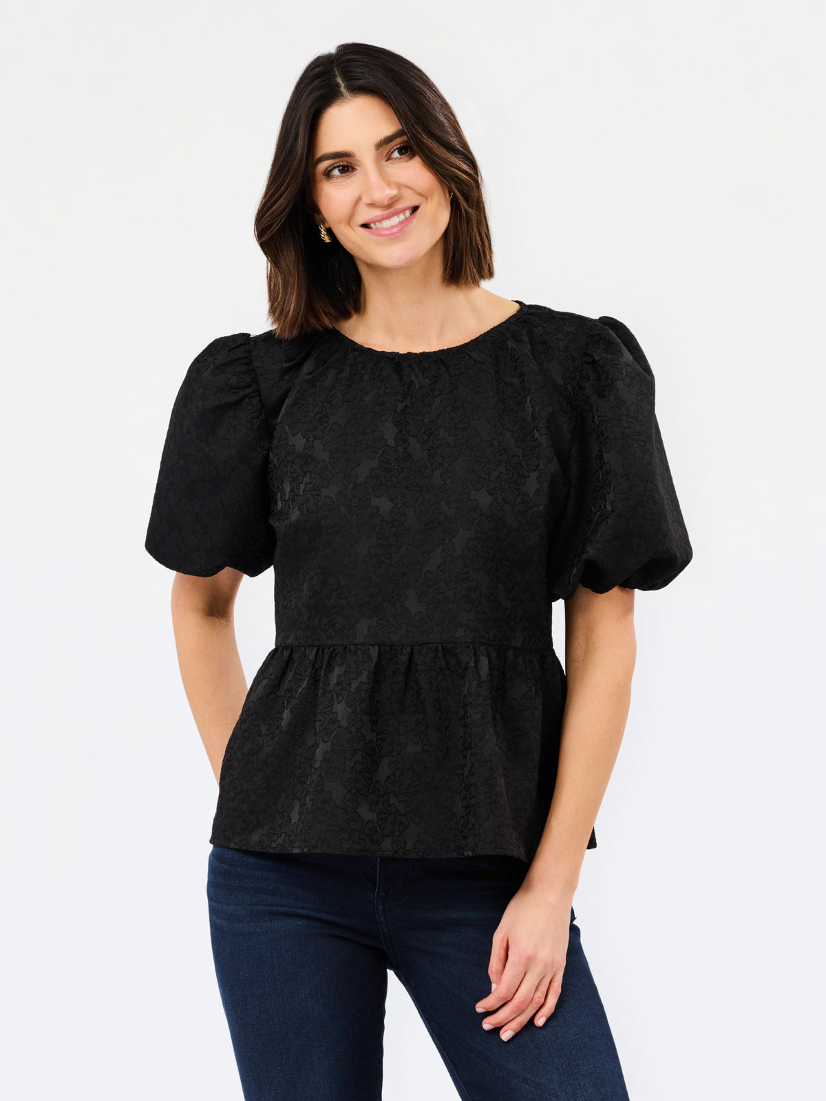 A woman modeling a black short-sleeve blouse with textured puff sleeves and a peplum waist, paired with dark blue jeans, against a plain white studio background.