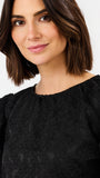 A close-up shot of a woman with a short brown bob haircut wearing a black textured long-sleeve top with a gathered neckline, set against a plain light gray background.