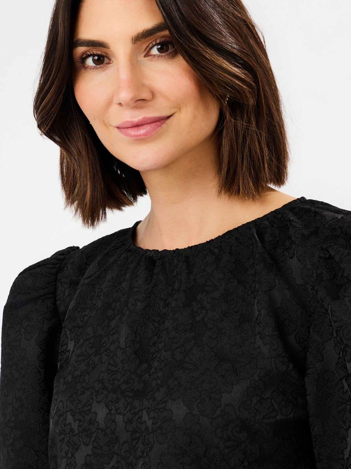 A close-up shot of a woman with a short brown bob haircut wearing a black textured long-sleeve top with a gathered neckline, set against a plain light gray background.