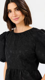 A woman wearing a black short-sleeve dress with textured jacquard fabric and puffed sleeves, shown against a plain light gray studio background.