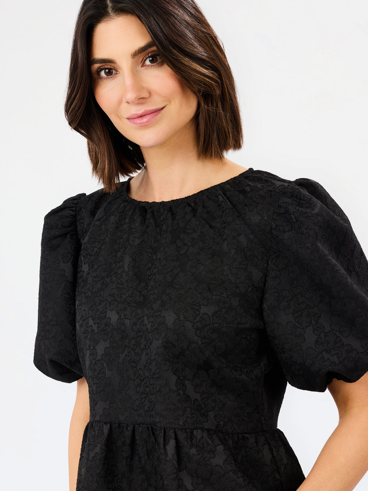 A woman wearing a black short-sleeve dress with textured jacquard fabric and puffed sleeves, shown against a plain light gray studio background.
