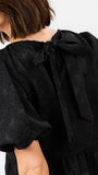 Close-up back view of a model wearing a black textured dress with a large bow at the back of the neck and puffed short sleeves.