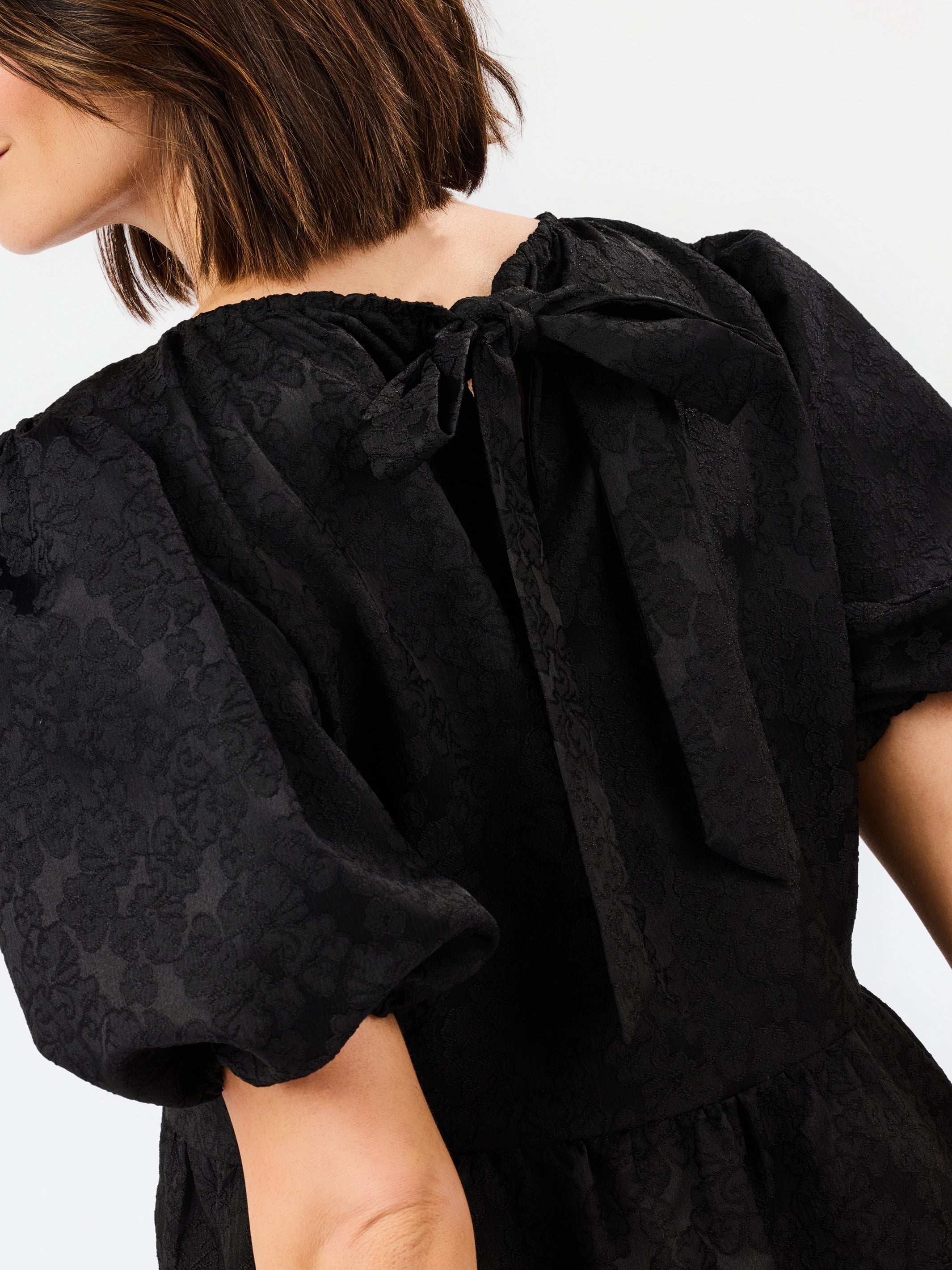Close-up back view of a model wearing a black textured dress with a large bow at the back of the neck and puffed short sleeves.