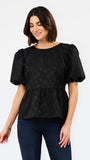 A woman wearing a black short-sleeve puff-sleeve peplum top with a subtle jacquard pattern, paired with dark blue jeans, against a plain white studio background.
