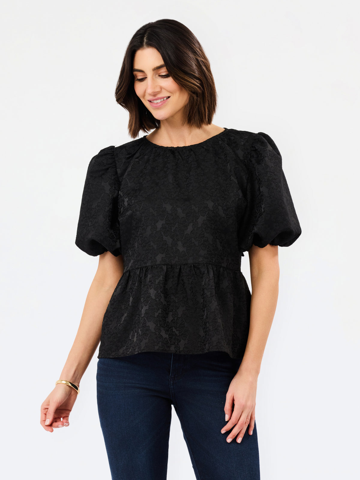 A woman wearing a black short-sleeve puff-sleeve peplum top with a subtle jacquard pattern, paired with dark blue jeans, against a plain white studio background.