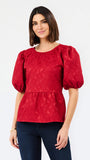 A woman modeling a red textured puff-sleeve blouse with a peplum waist, paired with dark blue jeans, against a plain white studio background.