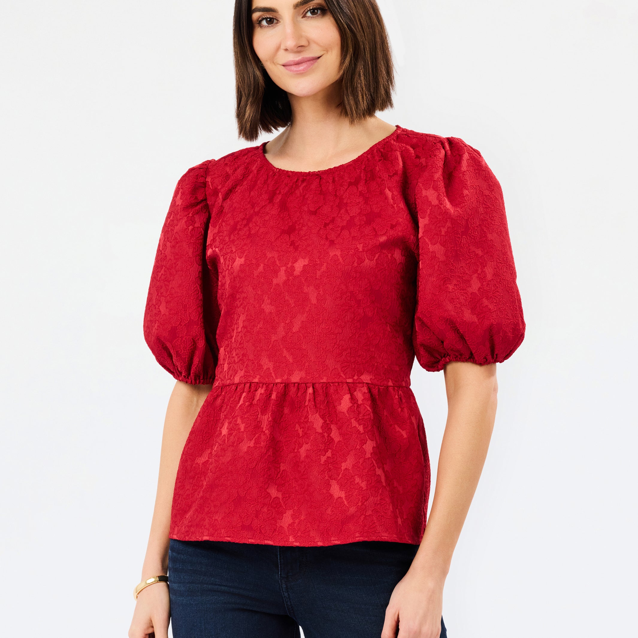 A woman modeling a red textured puff-sleeve blouse with a peplum waist, paired with dark blue jeans, against a plain white studio background.