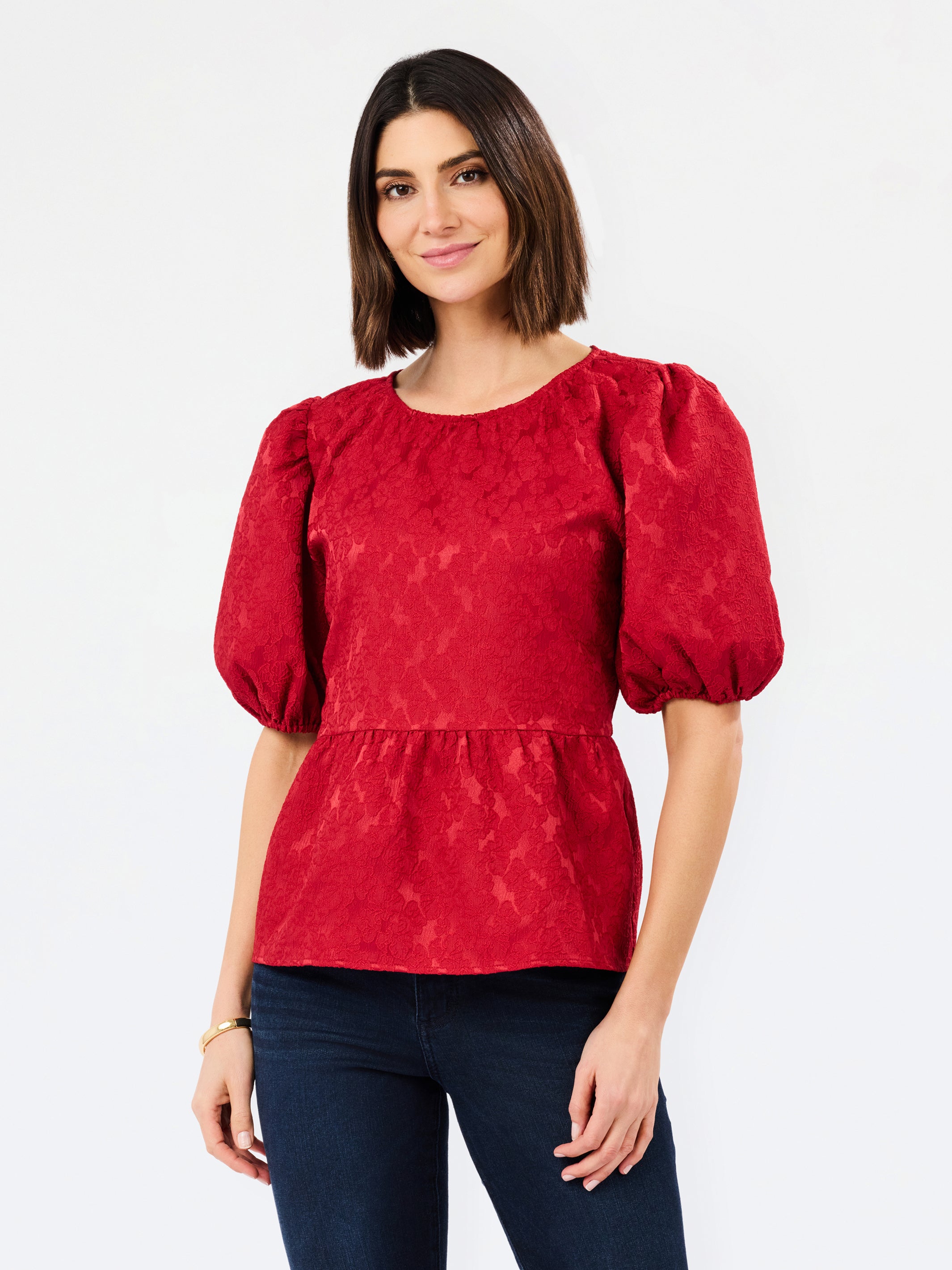 A woman modeling a red textured puff-sleeve blouse with a peplum waist, paired with dark blue jeans, against a plain white studio background.
