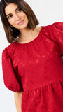 A woman wearing a red textured puff-sleeve top with a gathered waist, photographed against a plain light background.