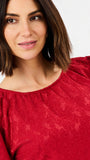 A close-up of a woman wearing a textured red top with star patterns and puffed sleeves, set against a plain light background.