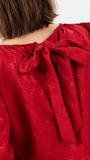 Close-up of the back of a red lace top with a large bow at the neckline, worn by a model with short brown hair against a plain light background.