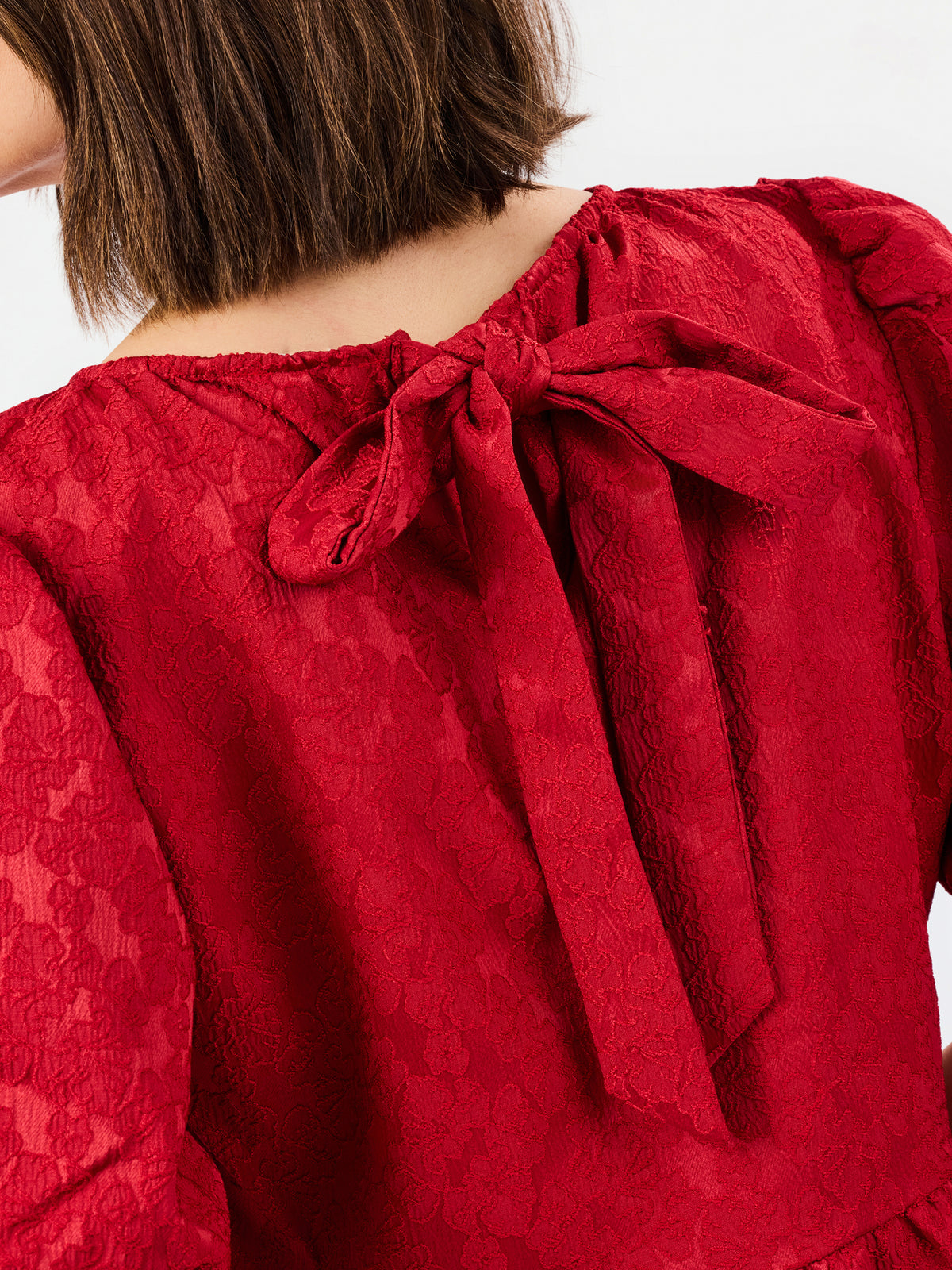 Close-up of the back of a red lace top with a large bow at the neckline, worn by a model with short brown hair against a plain light background.