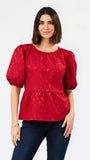 A woman modeling a red short-sleeve peplum blouse with puff sleeves and a textured, slightly mottled fabric, paired with dark blue jeans against a plain white background.